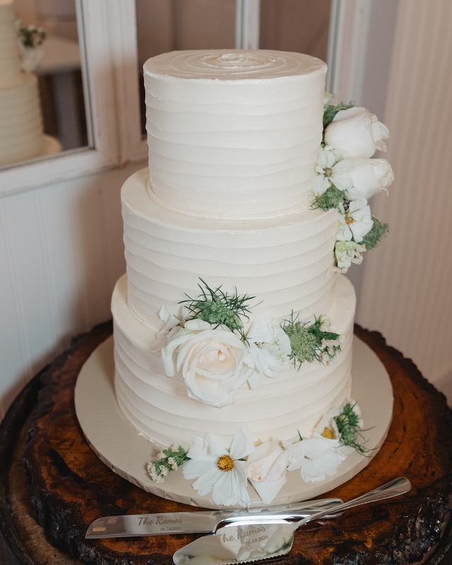 A three-tier white buttercream wedding cake with white roses and daisies, on a wood slab by a window with the cake knife and server