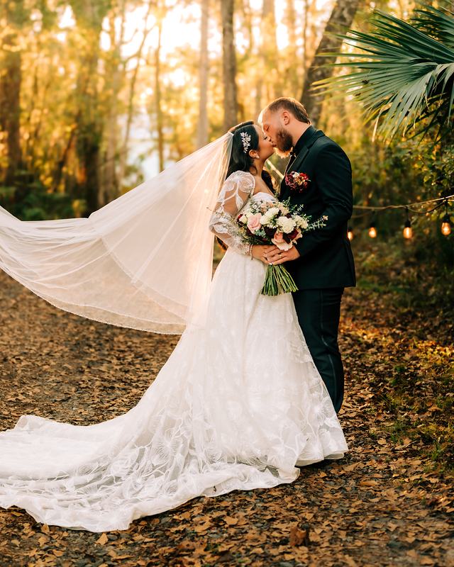 A bride’s long veil caught in the air as the couple kiss among the trees at golden hour