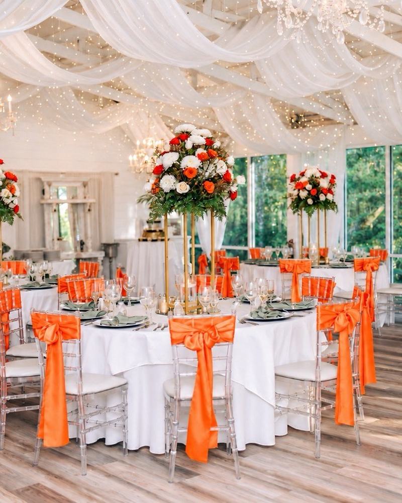 The Garden Room set for dinner: round tables in white linen with orange sashes on clear chairs, tall red and white floral centerpieces, white fabric swags and string lights overhead