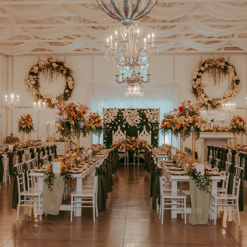 The Barn set for a reception: long white farm tables with sage runners and forest-green chair sashes, tall wildflower centerpieces, two floral wreaths and a white rose wall behind the sweetheart table
