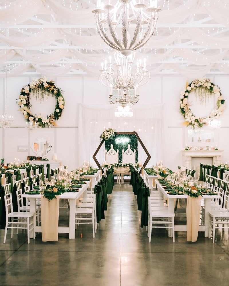 The Barn set for a reception: long white tables with dark green sashes and greenery runners, floral wreaths on the white walls and a hexagon arch at the head table, under crystal chandeliers