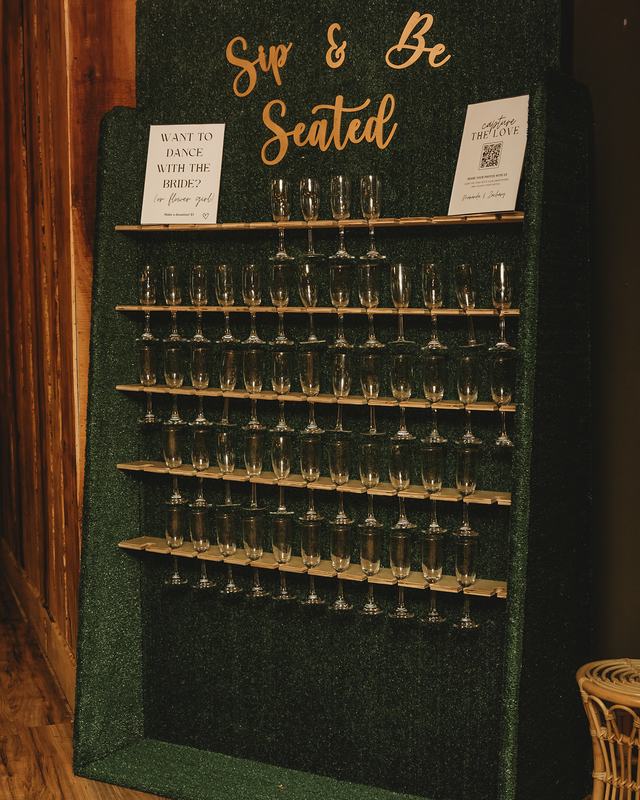 A dark green champagne wall lettered “Sip & Be Seated”, its shelves lined with filled flutes