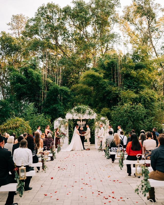 Guests seated either side of a petal-strewn stone aisle, the couple at a white garden arch beneath tall trees