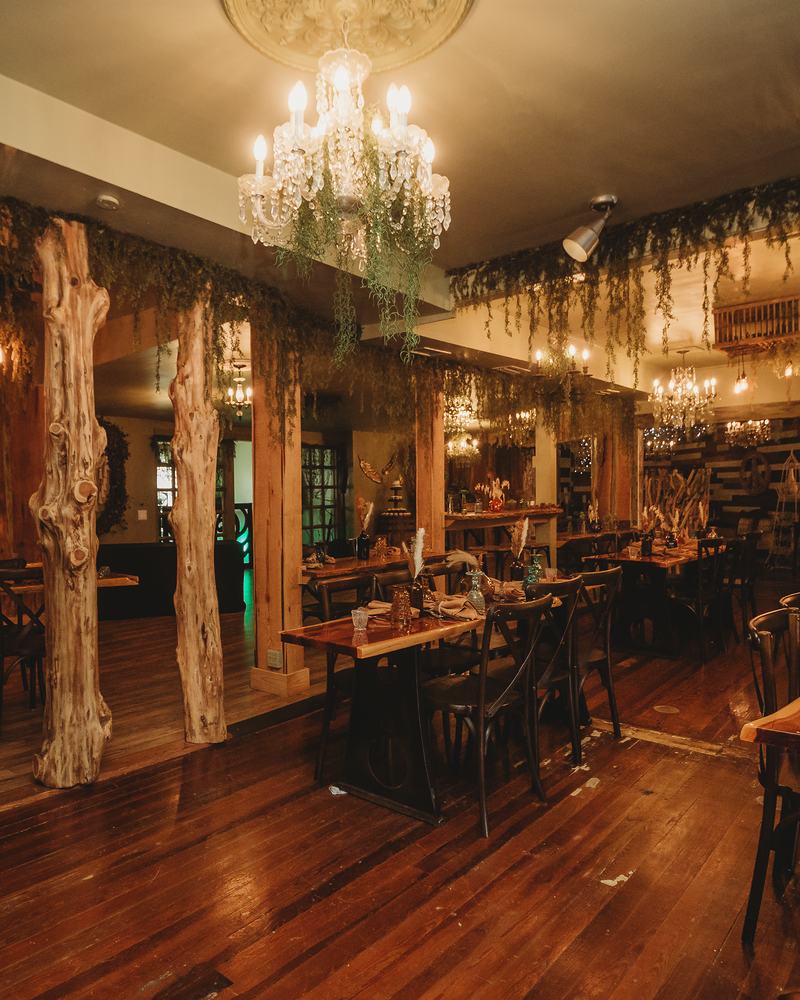 The BoHo dining room: live-edge tables and cross-back chairs between carved tree-trunk pillars, under crystal chandeliers and trailing greenery