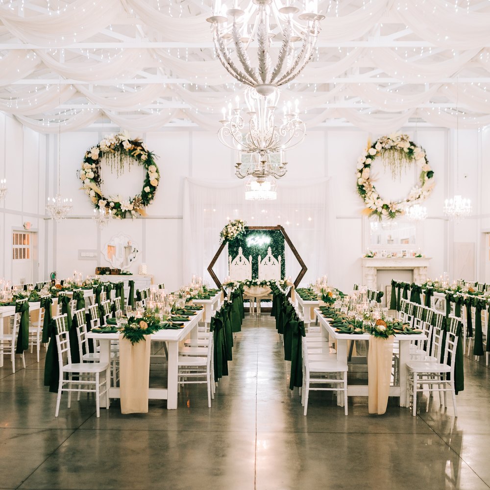 Long white farm tables inside the Barn, dressed with greenery runners and forest-green chair sashes beneath crystal chandeliers
