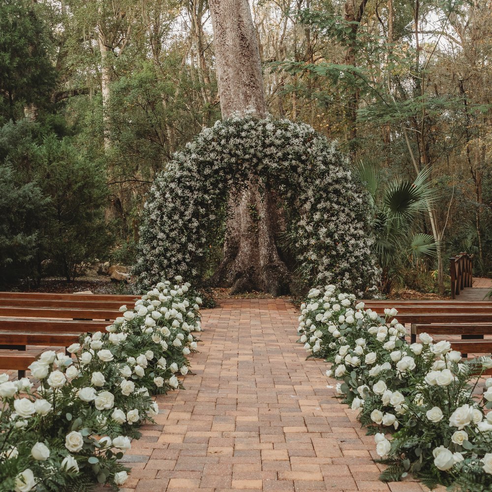 Ceremony aisle lined with white roses and wooden benches, leading to a flowering arch at the foot of an old oak