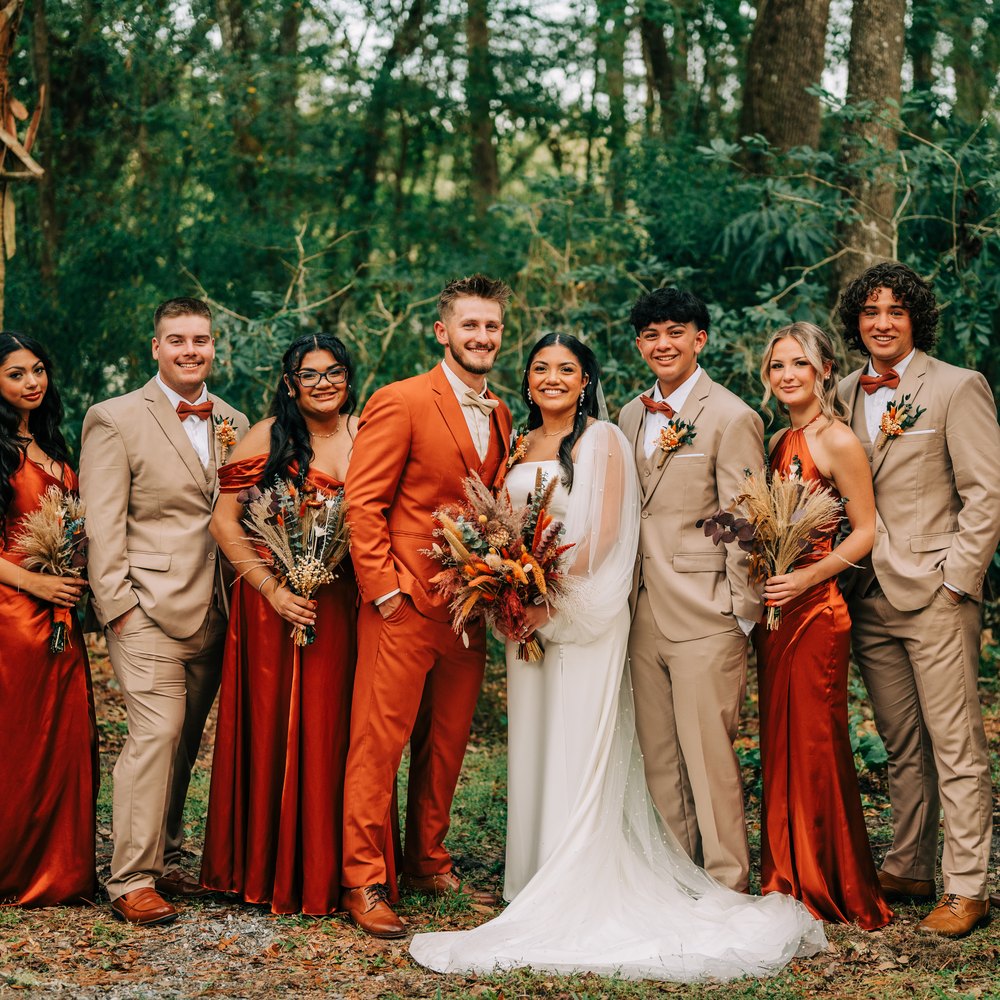 Bridal party in tan suits and rust satin dresses gathered around the couple in front of the woods