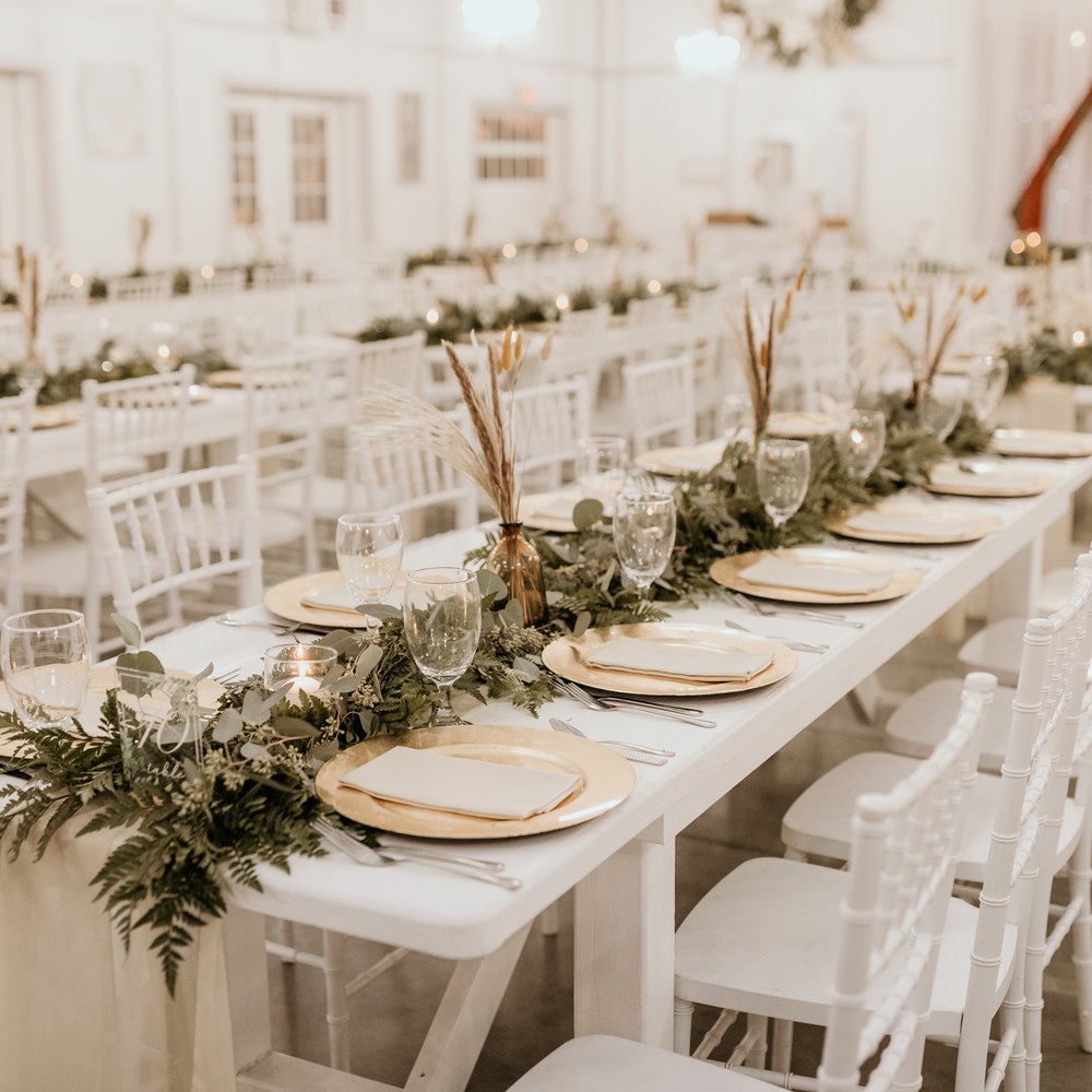Long white farm table in the Barn set with gold chargers, fern and eucalyptus garland and dried pampas, rows of white chairs beyond