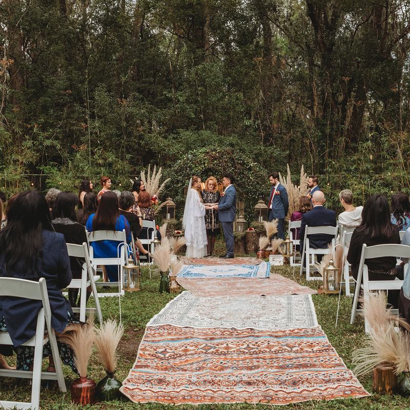An outdoor BoHo ceremony in the woods: layered rugs down the aisle, pampas grass along the chairs and guests seated either side