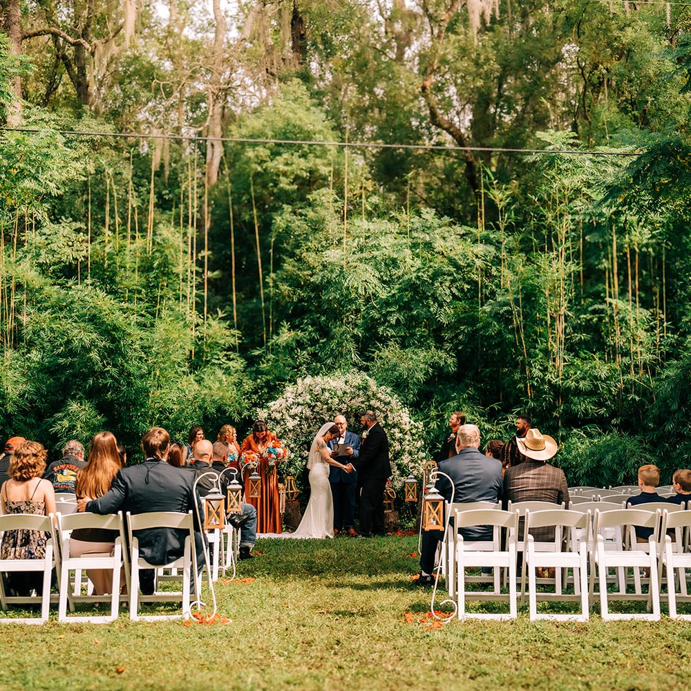 A ceremony in the woods beside the BoHo: guests on white chairs either side of a lantern-lined aisle, the couple at a flowering arch under tall bamboo
