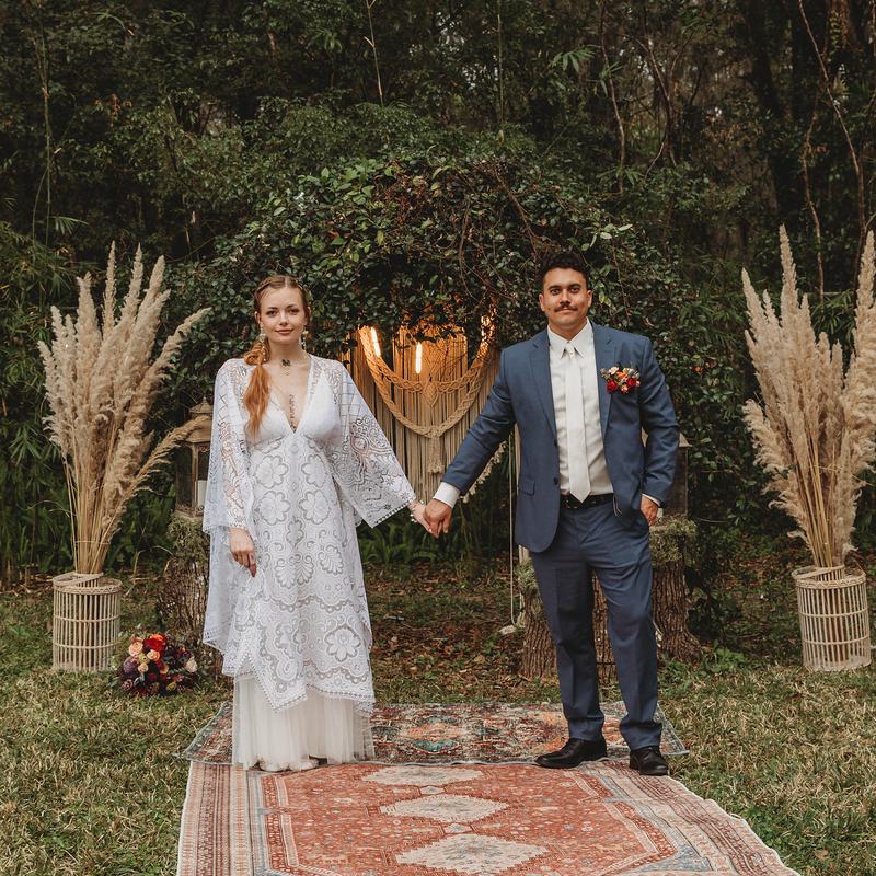 A bride in a lace boho gown and her groom holding hands on a rug between two urns of pampas grass, a macramé backdrop behind them