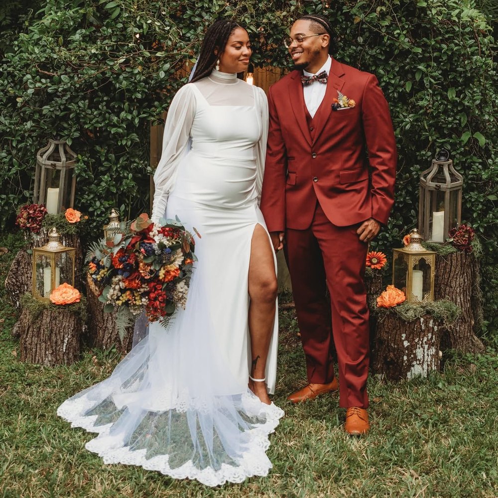 Bride and groom in front of the greenery wall outside the BoHo, lanterns and florals set on log stumps around them