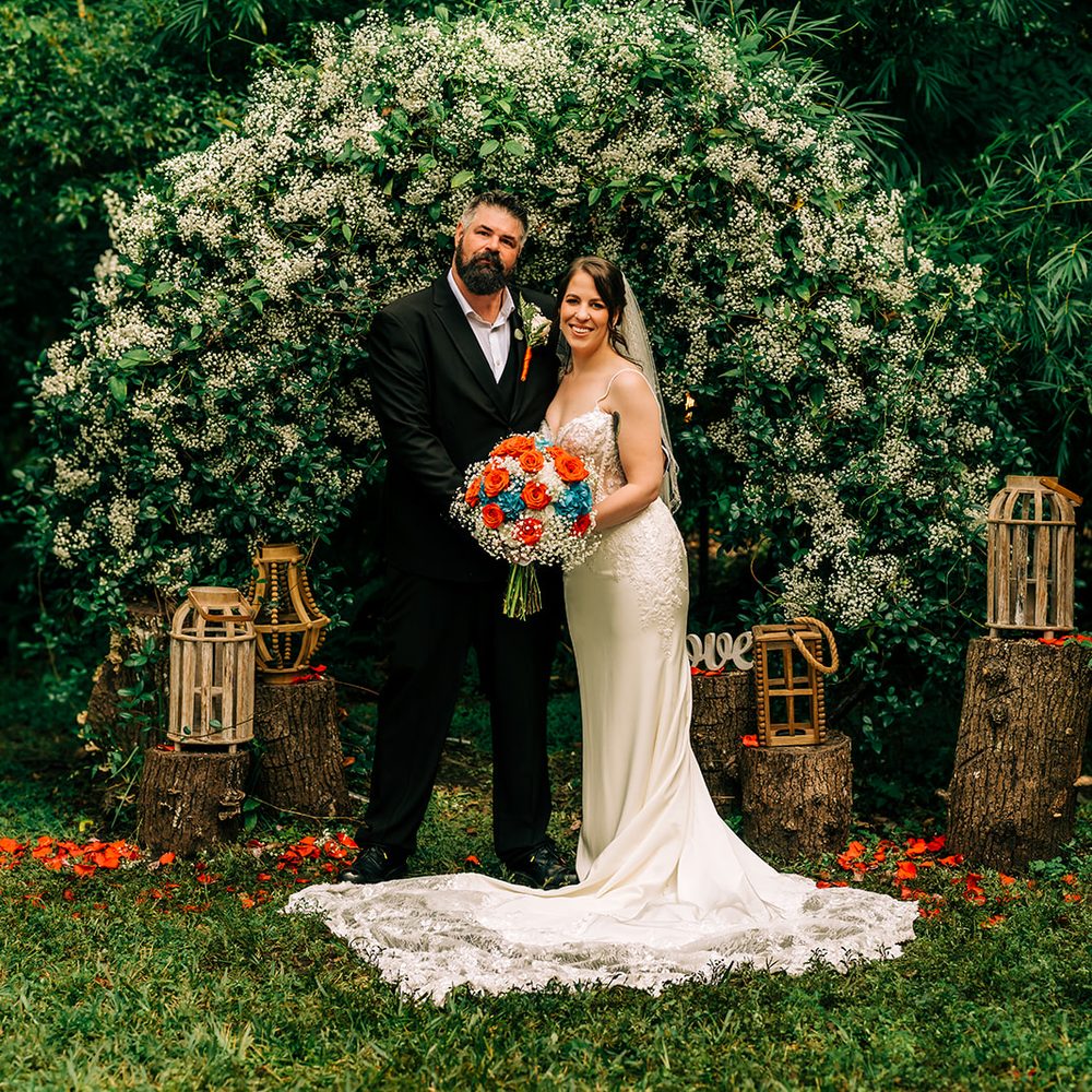 Bride and groom at the flowering arch in the BoHo woods, wooden lanterns on log stumps and orange petals scattered on the grass