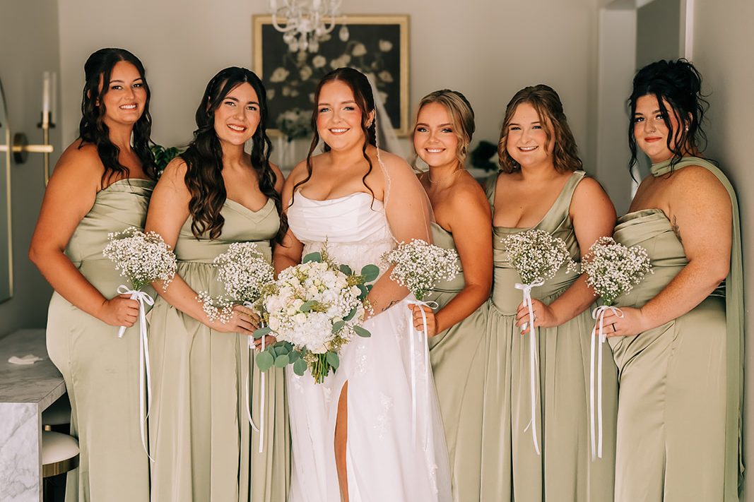 A bride and her five bridesmaids in sage satin gowns, holding baby's breath bouquets, inside the Bridal Suites