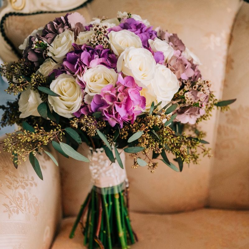 Bridal bouquet of purple hydrangea and ivory roses with seeded eucalyptus, resting on a cream armchair