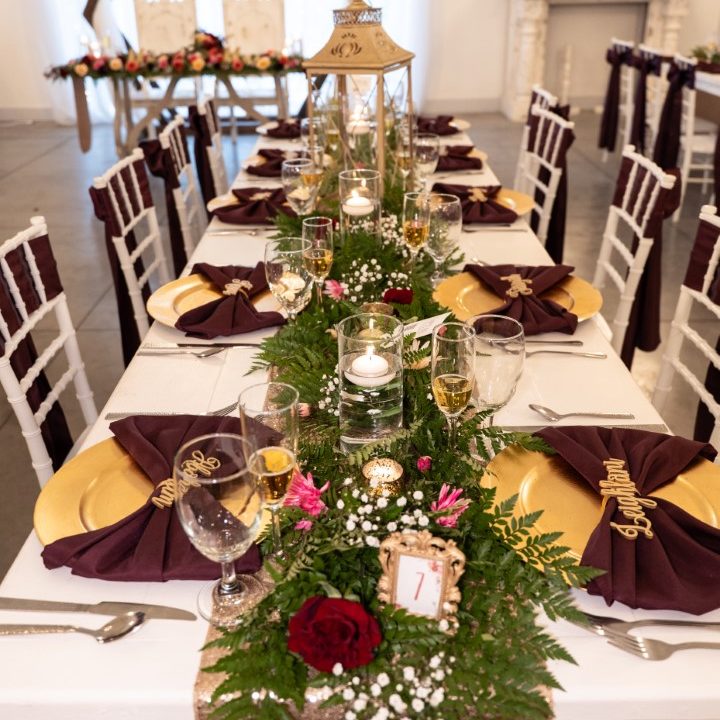Reception table runner of ferns, red roses and baby's breath between plum napkins and gold chargers