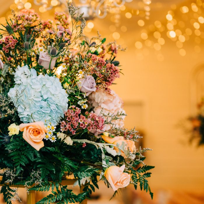 Floral arrangement of pale blue hydrangea, peach roses and wildflowers against warm string lights