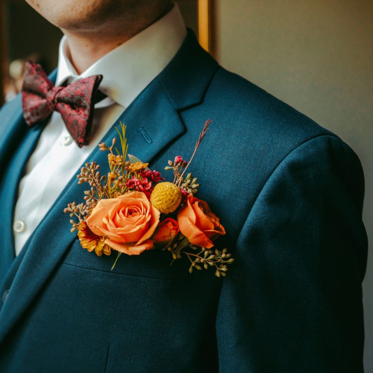 Orange rose and craspedia boutonniere on a teal suit lapel with a patterned bow tie