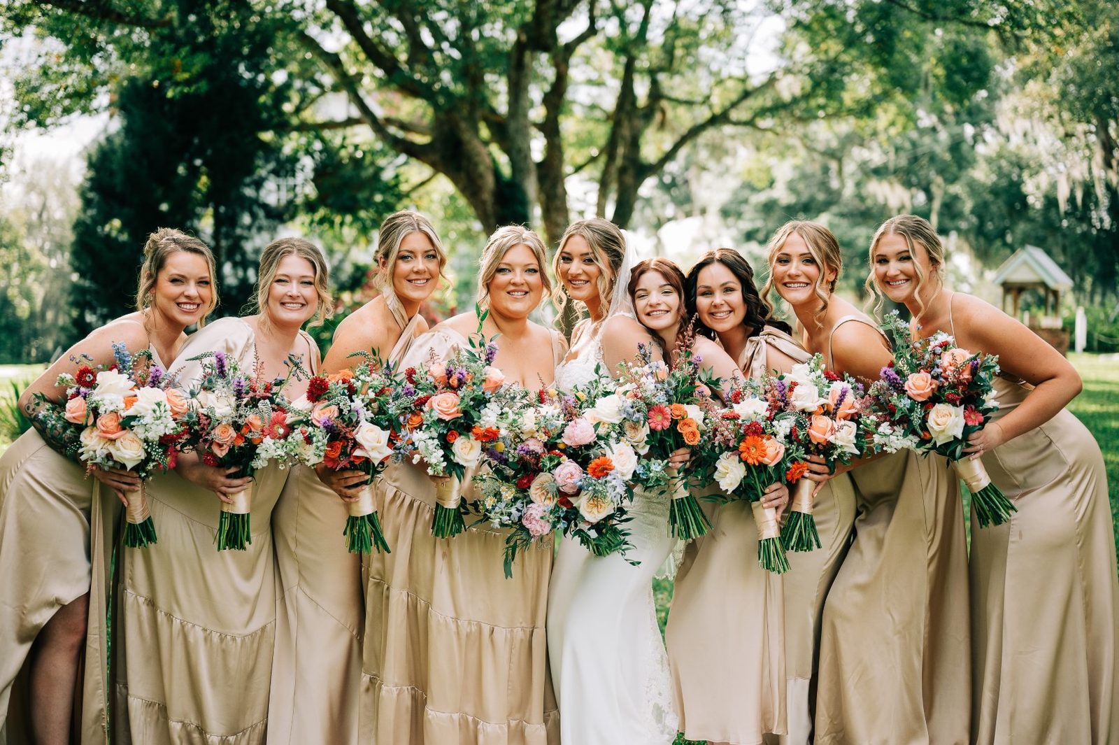 Six bridesmaids in champagne gowns holding colorful garden-style bouquets around the bride, under live oaks