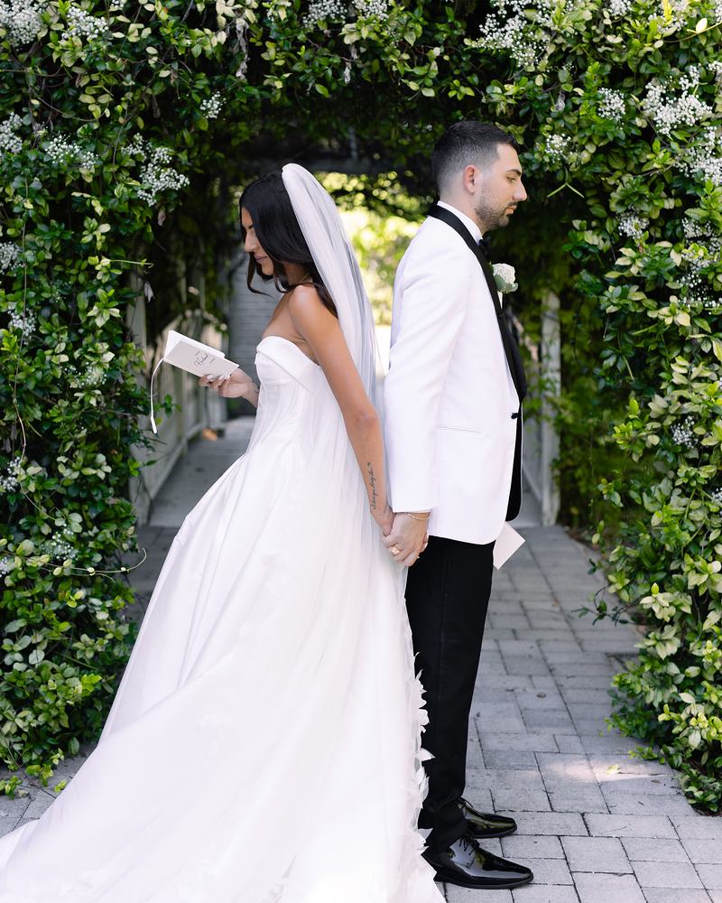 Bride and groom side by side beneath the jasmine-covered arbor on the brick path, the officiant reading from a small book