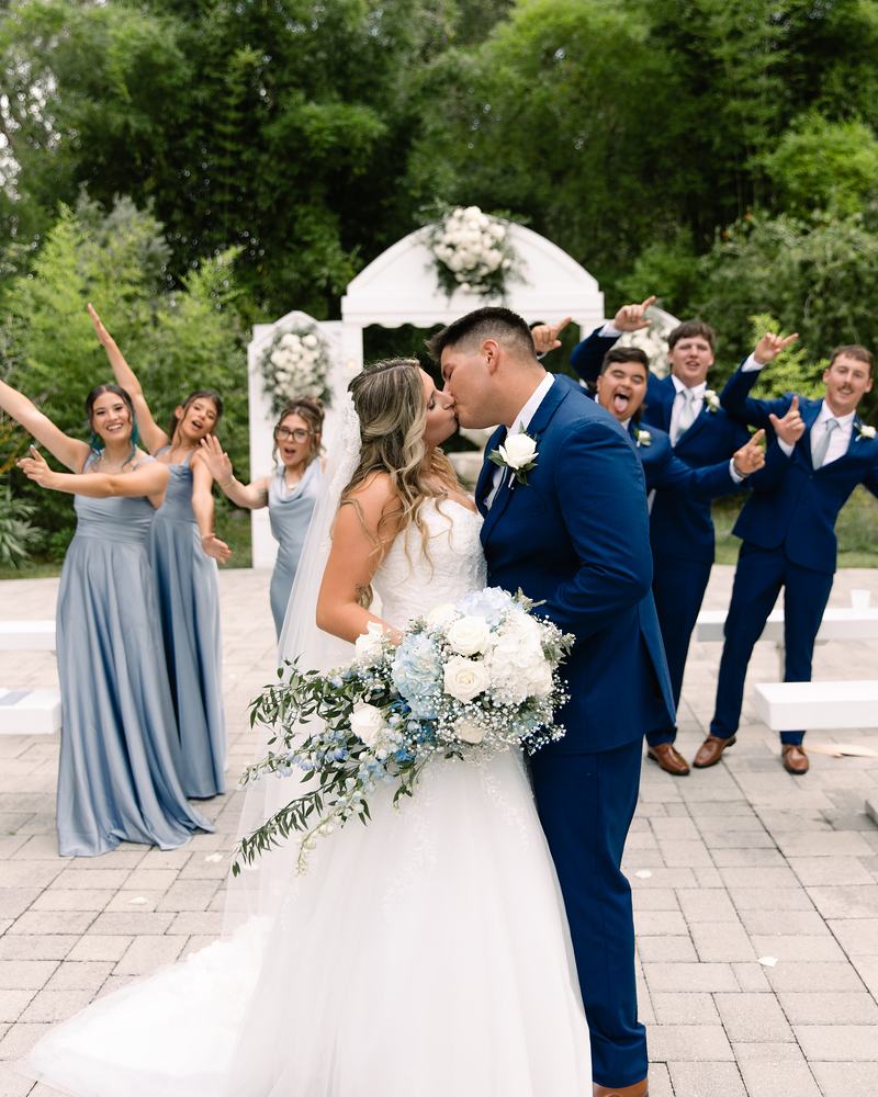 Bride and groom kissing at the white ceremony arch while bridesmaids in dusty blue and groomsmen in navy cheer behind them