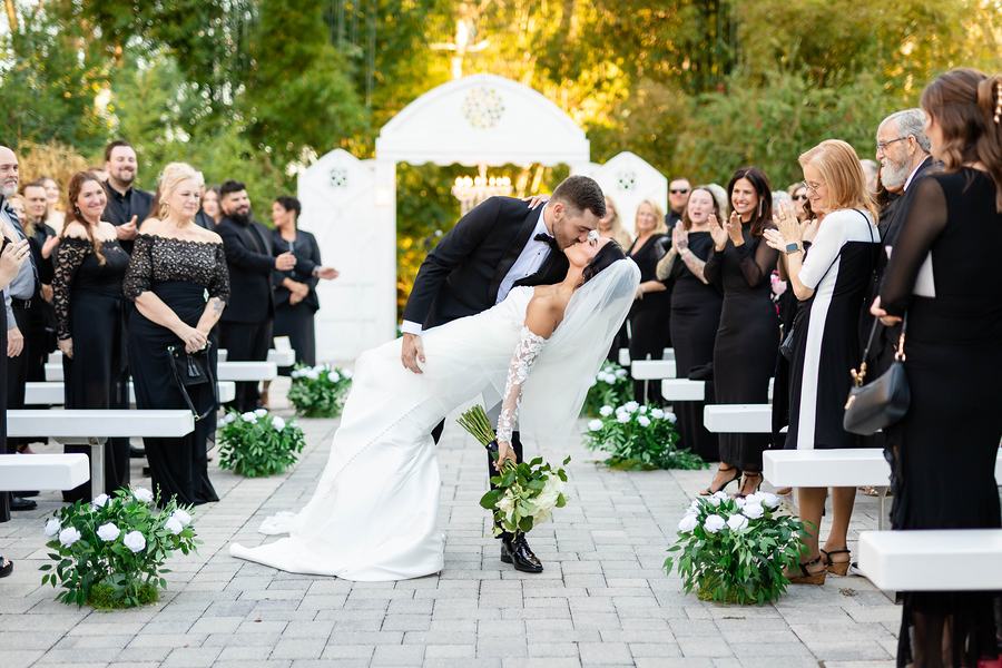 Groom dipping the bride for a kiss in the aisle of the garden ceremony, guests in black standing and cheering beside white benches