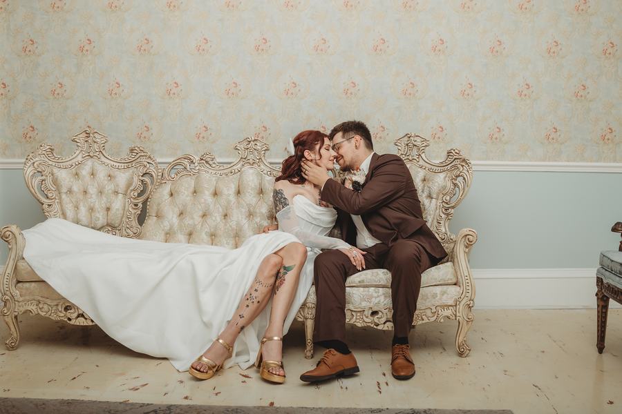 Bride and groom kissing on a cream tufted Victorian settee, her gown spread across the cushions and her boots on the floor