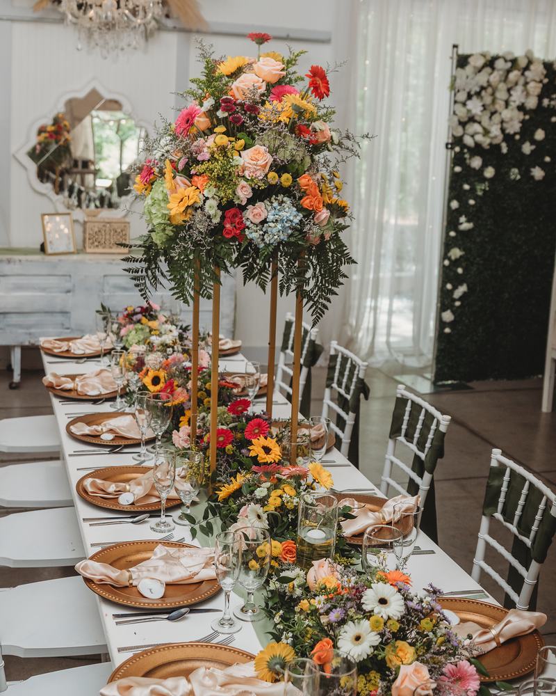 A long reception table set with gold chargers and folded napkins, tall arrangements of orange, yellow and pink flowers running down its center