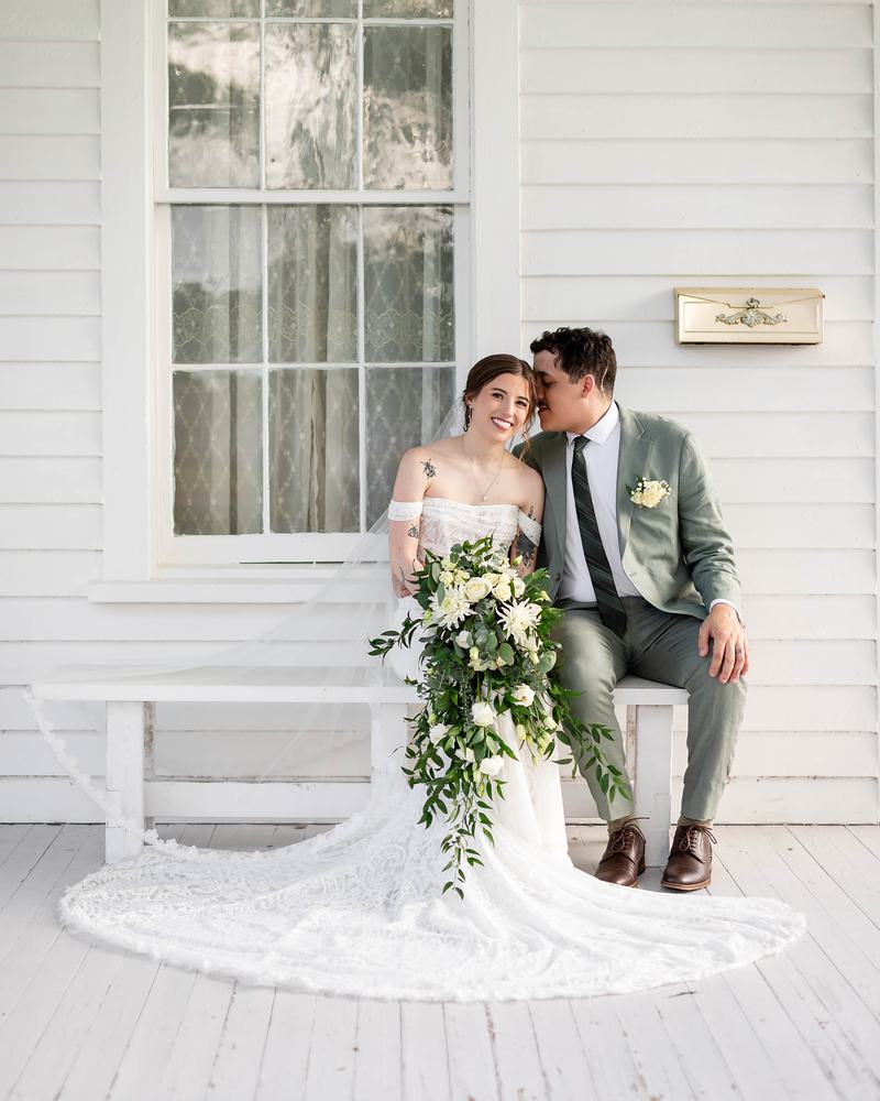 Bride and groom sitting close on a white bench against the white clapboard wall, her cascading bouquet across her lap