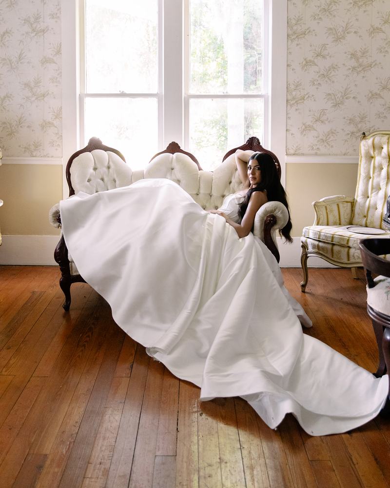 Bride reclining on a carved white settee by the window, her train flowing across the wooden floor
