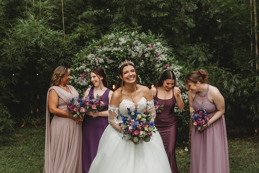 Bride laughing with her bridesmaids in shades of plum and mauve, flowering shrubs behind them