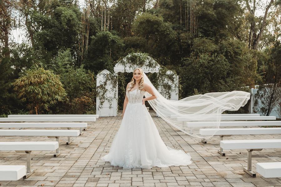 Bride standing on the ceremony lawn's stone path, her veil lifted sideways by the wind, the white arch and benches behind her