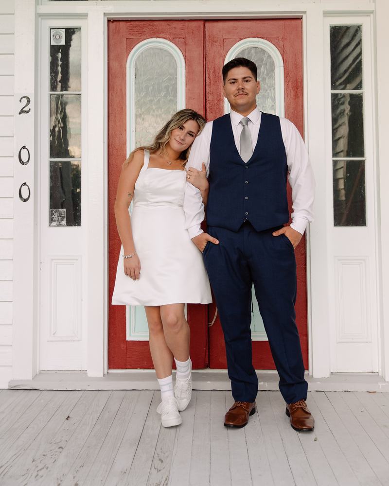 Bride and groom leaning in the doorway of the red double doors, she in a short white dress and he in a navy waistcoat