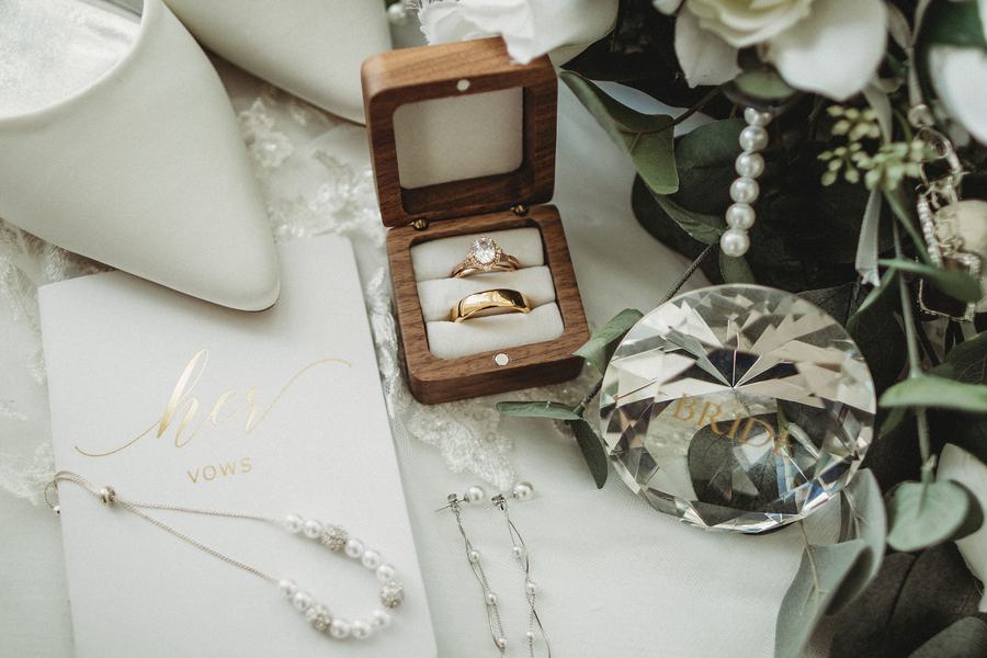 Wedding rings in a wooden box beside her vow book, white shoes, pearls and greenery laid out on lace