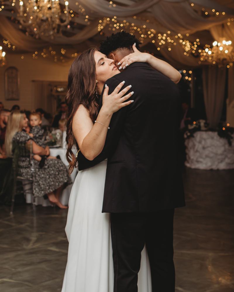 Bride and groom holding each other close for a first dance beneath the chandelier and string lights