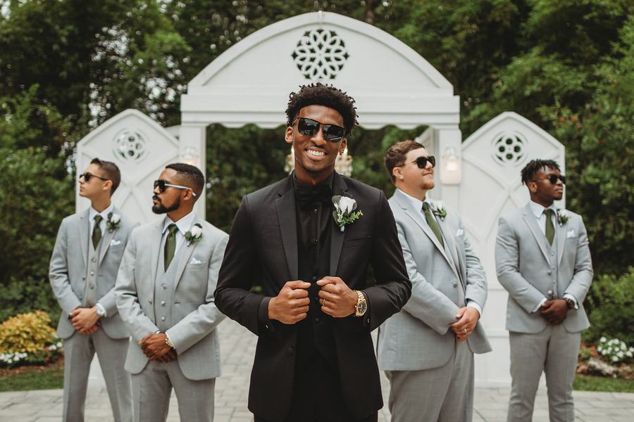 Groom in a black tuxedo grinning in front of the white arch, his groomsmen in gray suits and sunglasses either side
