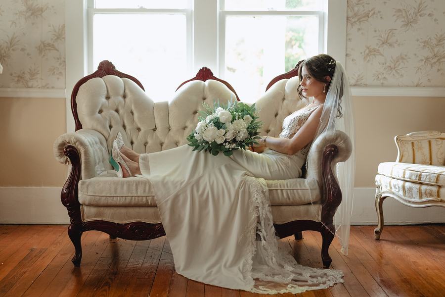 Bride seated on a carved settee by the window, her bouquet resting beside her and her veil falling to the floor