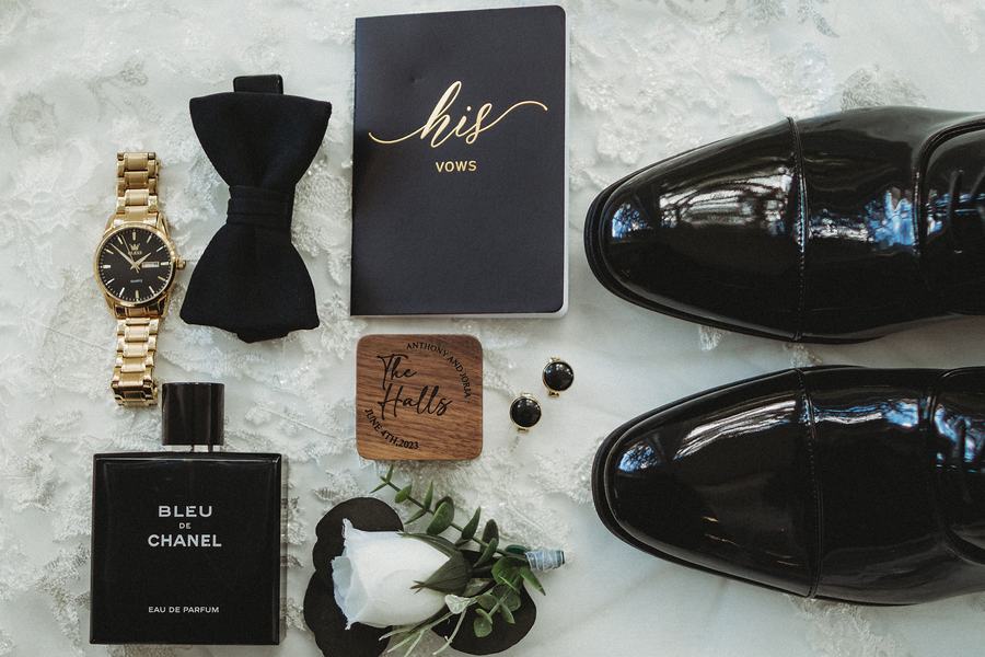 Groom's details laid out on lace: his vow book, black bow tie, watch, cologne, cufflinks and patent shoes
