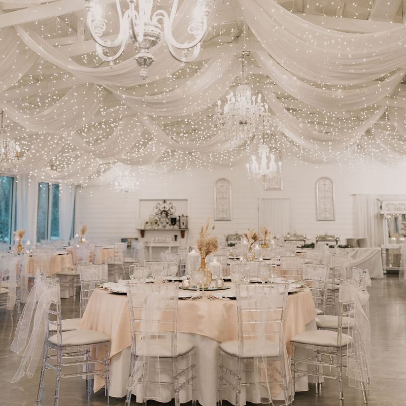 The Garden Room set for a reception: round tables in ivory linen and clear chairs beneath swags of white fabric, fairy lights and crystal chandeliers