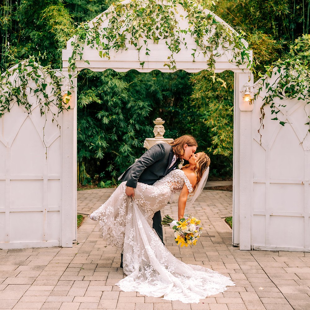 Groom dipping the bride for a kiss beneath the white garden arbor, its doors trailing with ivy