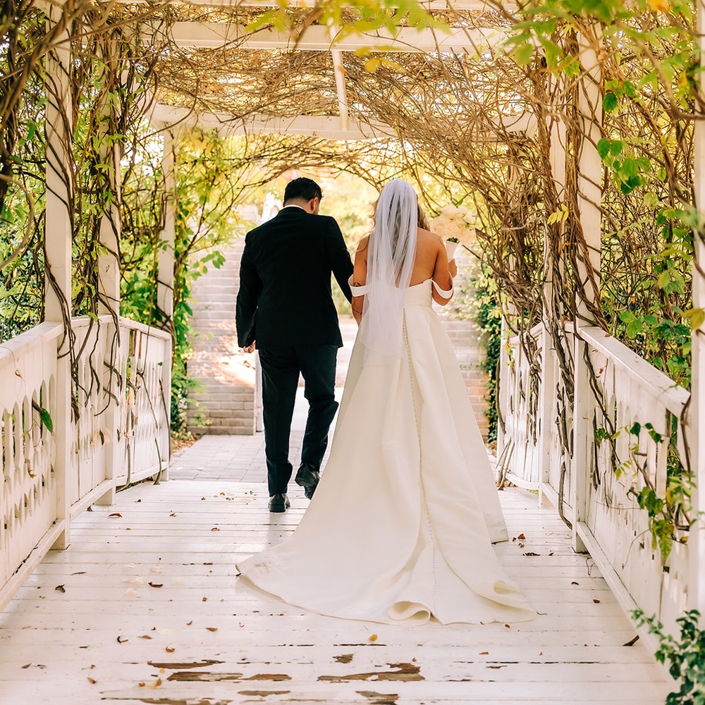 Bride and groom walking hand in hand down the vine-covered pergola in the gardens