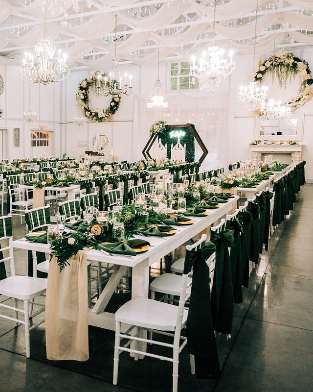 The Barn set for dinner: long white tables with dark green napkins and greenery runners beneath crystal chandeliers