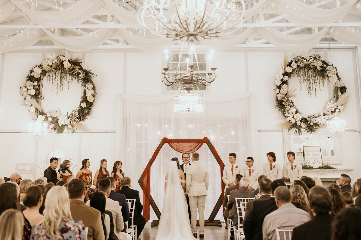A ceremony held indoors in the Barn: guests seated facing a copper hexagon arch, floral wreaths and crystal chandeliers overhead